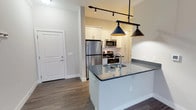 Located at 1221 E Broadway in Louisville, KY, this single-floor unit offers a thoughtfully designed layout with modern finishes throughout. The open-concept kitchen serves as the heart of the space, showcasing white shaker cabinetry, a subway tile backsplash, stainless steel appliances including a refrigerator, electric range, and microwave, and a center island with dark granite-style countertops and an undermount sink. Stylish pendant lights hang above the island, complemented by track lighting overhead, creating a well-lit and functional cooking and entertaining area. Rich wood-look flooring extends throughout the main living areas, adding warmth and continuity. The kitchen connects seamlessly to the living area, which features french doors that invite natural light and provide access to an outdoor space. A dedicated laundry area is conveniently tucked just off the kitchen. The bathroom is elegantly appointed with large-format marble-look tile in the walk-in shower, a white vanity with stone countertops, and matching wood-look flooring. The bedroom is generously proportioned, offering plush carpet, a ceiling fan, and two windows that fill the room with natural light, creating a comfortable and serene retreat.