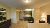 This single-floor studio unit offers an efficient, open-concept layout that integrates living, dining, and sleeping areas into one cohesive space. The flooring throughout is light hardwood, providing a warm and clean aesthetic that ties the areas together seamlessly.

The kitchenette is nestled within a recessed nook and features dark cabinetry with ample upper and lower storage, a countertop with a built-in sink, and under-cabinet lighting that brightens the workspace. A black microwave and additional appliances are included, making the kitchen fully functional. Adjacent to the kitchenette, a compact dining area accommodates a small table with chairs, ideal for everyday meals.

The main living and sleeping area is generously proportioned relative to the overall footprint, with neutral-toned walls and a textured ceiling that add subtle dimension to the space. A hallway connects the main area to additional storage, including sliding-door closets that offer practical organization solutions.

Flush-mount ceiling fixtures provide consistent overhead lighting throughout. The overall design is straightforward and functional, with a neutral color palette of warm beige and green tones that creates a comfortable, livable environment suited for individuals seeking an efficient yet well-appointed space.