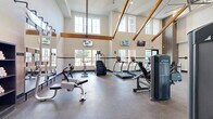 This fitness center is a comprehensive amenity space spread across three floors, with the primary exercise areas occupying the main level. The facility is architecturally distinguished by soaring vaulted ceilings with exposed wooden beam trusses, generous wall-to-wall windows, and durable rubber flooring throughout — all contributing to a bright, open, and energizing workout environment.

The main exercise floor is generously sized and houses a robust selection of True-brand cardio equipment, including multiple treadmills, stair climbers, and upright cycles, as well as a rowing machine for full-body conditioning. A dedicated strength training zone features a multi-station cable machine, a dumbbell rack, and various free weight accessories. Wall-mounted televisions are positioned throughout for entertainment during workouts. A smaller, secondary exercise area on an upper level provides additional space for focused training. Towel service and storage cubbies are available near the entrance for added convenience.

Beyond the fitness center, the surrounding outdoor amenity areas are equally impressive. A resort-style swimming pool with an expansive sun deck, lounge chairs, and shaded seating areas serves as a central outdoor gathering space. Landscaped courtyards with mature trees, concrete pathways, and outdoor dining furniture provide attractive, well-maintained common areas. The building exterior blends brick, glass, and modern panel finishes, creating a contemporary yet welcoming community environment.