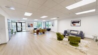 Located at 900 Farmington Ave in West Hartford, Connecticut, this thoughtfully designed amenity space offers a versatile and stylish environment across a single floor. Light hardwood flooring and a clean white drop-tile ceiling with recessed panel lighting run throughout, creating a bright, cohesive aesthetic.

The main lounge area is generously proportioned and filled with natural light from large floor-to-ceiling windows overlooking the surrounding neighborhood. It features a comfortable seating arrangement with a gray sofa, green velvet accent chairs, and a curated area rug, alongside wall-mounted artwork. A large-screen TV is mounted on the wall, making this area ideal for relaxing or casual gatherings. Plush white boucle lounge chairs and a second seating vignette add to the social atmosphere, complemented by a shuffleboard table for recreation.

A dedicated conference room provides a professional setting with a long light wood table, ergonomic rolling chairs, a wall-mounted display screen, and built-in power access — well-suited for meetings or presentations. An adjacent row of green stackable chairs offers additional flexible seating.

The kitchenette features white cabinetry, a stainless steel refrigerator, microwave, and a blue tile backsplash, providing practical support for gatherings. Together, these spaces form a well-rounded, modern communal environment.