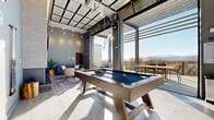 This contemporary rooftop amenity space seamlessly blends indoor comfort with outdoor recreation, all set against breathtaking panoramic mountain views. The centerpiece of the indoor area is a stylish pool table positioned beneath a retractable glass ceiling, which opens directly onto the outdoor terrace — flooding the space with natural light and creating a fluid indoor-outdoor experience. Large floor-to-ceiling glass panels and industrial-style overhead doors further enhance the open, airy atmosphere.

The lounge area features a sleek linear fireplace set into a dramatic floor-to-ceiling textured tile accent wall, accompanied by modern furnishings including a marble-top coffee table, plush seating, and curated décor. Pool cues are neatly mounted on the wall nearby, underscoring the space's focus on recreation.

The fully equipped kitchen is appointed with warm wood cabinetry, quartz countertops, a large island with bar seating, stainless steel appliances including a full-size refrigerator, and a professional-grade range hood — ideal for entertaining.

The expansive outdoor terrace offers a foosball table, bench seating, a gas grill station, and open-air dining, all framed by cable-rail fencing and a pergola overhead. Sweeping mountain and cityscape views make this terrace a standout feature of the entire space.