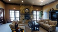 This single-floor space exudes a refined, lodge-inspired ambiance that balances rustic charm with classic elegance. The centerpiece of the main lounge area is a striking floor-to-ceiling natural stone fireplace, crowned with a decorative round mirror and flanked by carefully curated accents that add depth and character to the room. Recessed lighting complements the warm earth-toned walls, creating a cozy yet polished atmosphere throughout. Large French-style windows and doors line the walls on either side of the fireplace, flooding the space with generous natural light and offering views to the exterior. The seating arrangement features plush, neutral-toned sofas and accent chairs positioned around a dark wood coffee table, creating an intimate and functional social setting. Rich dark wood trim frames the windows, doorways, and ceiling line, lending architectural definition and a sense of sophistication. Decorative elements, including ornate vases, candleholders, and lush indoor greenery, are thoughtfully placed to enhance the space's inviting character. The carpeted flooring and warm color palette throughout contribute to an overall sense of comfort and warmth, making this a welcoming environment well-suited for relaxed gatherings and socializing.