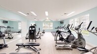 Located at Marley Manor in Salisbury, Maryland, this dedicated fitness center offers a comprehensive workout environment designed to support a wide range of exercise routines. The single-floor facility is finished with light wood-look flooring and soft aqua-toned walls, creating a clean, airy atmosphere throughout.

The space is generously lit by a combination of recessed panel lighting and skylights, while multiple windows along the walls invite natural light and provide views of the surrounding community. Vaulted ceiling sections in key areas of the room add a sense of openness and volume.

The cardio section is well-equipped with a row of treadmills, elliptical trainers, and a rowing machine, all positioned to take advantage of the natural light streaming through the windows. A wall-mounted television is conveniently placed for entertainment during workouts.

The strength training area features a multi-station cable machine, a cable crossover unit, a dumbbell rack stocked with a full range of free weights, and multiple adjustable workout benches. Full-length mirrors line one wall, enhancing the sense of space and allowing users to monitor their form.

A dedicated open floor area with built-in storage cubbies provides space for stretching, mat work, or group fitness activities. The facility also includes security surveillance and is accessible via double doors, rounding out a functional and well-maintained fitness amenity.