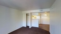 This single-floor unit offers a clean, functional layout designed to maximize everyday living. The main living area is finished with plush dark carpeting and features a built-in sliding-door closet, providing convenient and accessible storage. Neutral white walls and a smooth ceiling create a bright, airy atmosphere throughout the space.

The living area flows seamlessly into the kitchen, which is defined by a peninsula-style counter that serves as both a workspace and a casual dining surface. The kitchen is equipped with light-toned cabinetry offering ample storage, a refrigerator, and overhead ventilation. A pendant light fixture hangs above, adding a warm focal point to the cooking area.

Adjacent to the kitchen, a separate room is visible through an open doorway, illuminated by warm lighting, suggesting a dedicated dining or secondary living space. The hard flooring in the kitchen and adjoining room contrasts with the carpeted main area, helping to visually define each zone.

Central air distribution is provided via ceiling vents, ensuring consistent climate control throughout. The overall design emphasizes practicality and ease of use, with an efficient, well-connected floor plan that suits a range of lifestyles.