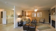 This single-floor space features an open-concept layout that connects the living area to the kitchen, creating a seamless flow throughout. The living room is generously sized, accommodating a full seating arrangement that includes a sofa, accent chairs, and leather ottomans centered around a dark-finished coffee table. Warm-toned walls in a soft amber hue contribute to the inviting ambiance, complemented by neutral carpet flooring accented with a striped area rug that helps define the seating zone. A ceiling fan with an integrated light fixture is centrally positioned overhead, providing both air circulation and ambient lighting. Table lamps on coordinating dark end tables add layered lighting throughout the room. Framed artwork adorns the walls, adding a decorative touch that enhances the overall aesthetic. A structural column near the kitchen entryway subtly separates the two spaces while maintaining visual openness. The kitchen, visible from the living area, features light cabinetry and a bright overhead light, offering functional workspace. Overall, the space presents a balanced combination of warmth, practicality, and comfortable design.