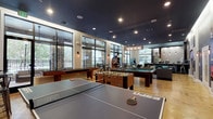This single-floor amenity space serves as a dynamic community hub designed for recreation and socializing. The main game room is a standout feature, offering an impressive lineup of entertainment options including a billiards table, foosball table, ping pong table, shuffleboard, and a classic arcade machine. The polished concrete floors, adorned with a subtle graphic design, complement the striking dark navy ceiling outfitted with recessed and pendant lighting that creates a vibrant yet inviting atmosphere. Large floor-to-ceiling windows and glass doors line the perimeter, flooding the space with natural light and providing views of the surrounding outdoor greenery and balcony areas. Stone accent columns add an earthy, architectural contrast to the modern interior palette. A dedicated lounge zone offers plush leather sofas and armchairs arranged around an area rug, alongside wall-mounted televisions, creating a comfortable setting for relaxation. Movie posters and decorative wall art add character to the interior. A separate, sleek kitchenette area features stainless steel cabinetry, a light countertop, pendant lighting, and a polished concrete floor, providing a functional service space. Together, these zones form a well-rounded communal environment designed for both active entertainment and casual gathering.