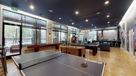 This single-floor amenity space serves as a dynamic community hub designed for recreation and socializing. The main game room is a standout feature, offering an impressive lineup of entertainment options including a billiards table, foosball table, ping pong table, shuffleboard, and a classic arcade machine. The polished concrete floors, adorned with a subtle graphic design, complement the striking dark navy ceiling outfitted with recessed and pendant lighting that creates a vibrant yet inviting atmosphere. Large floor-to-ceiling windows and glass doors line the perimeter, flooding the space with natural light and providing views of the surrounding outdoor greenery and balcony areas. Stone accent columns add an earthy, architectural contrast to the modern interior palette. A dedicated lounge zone offers plush leather sofas and armchairs arranged around an area rug, alongside wall-mounted televisions, creating a comfortable setting for relaxation. Movie posters and decorative wall art add character to the interior. A separate, sleek kitchenette area features stainless steel cabinetry, a light countertop, pendant lighting, and a polished concrete floor, providing a functional service space. Together, these zones form a well-rounded communal environment designed for both active entertainment and casual gathering.