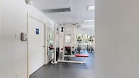 This fitness center offers a well-organized and fully equipped workout space designed to support a wide range of exercise routines. The room features durable dark rubber flooring throughout, providing a safe and functional surface for all types of training activities. Large windows and glass-paneled doors line the walls, flooding the space with natural light while offering pleasant views of the surrounding outdoor landscape and mature trees.

The strength training area is outfitted with multiple cable and pulley machines, including a multi-station cable system and a functional trainer, as well as adjustable benches in both flat and incline configurations. Full-length wall mirrors enhance the sense of openness and allow users to monitor their form during workouts. The cardio section features a treadmill, recumbent bike, and elliptical machines, all positioned near the large windows to make the most of the outdoor views. A wall-mounted television is also present for entertainment during cardio sessions.

The facility includes an accessible restroom, ceiling-mounted ventilation units for climate control, and recessed lighting to ensure a bright and comfortable environment. The clean white walls and organized layout contribute to a bright, motivating atmosphere well-suited for daily fitness use.