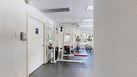 This fitness center offers a well-organized and fully equipped workout space designed to support a wide range of exercise routines. The room features durable dark rubber flooring throughout, providing a safe and functional surface for all types of training activities. Large windows and glass-paneled doors line the walls, flooding the space with natural light while offering pleasant views of the surrounding outdoor landscape and mature trees.

The strength training area is outfitted with multiple cable and pulley machines, including a multi-station cable system and a functional trainer, as well as adjustable benches in both flat and incline configurations. Full-length wall mirrors enhance the sense of openness and allow users to monitor their form during workouts. The cardio section features a treadmill, recumbent bike, and elliptical machines, all positioned near the large windows to make the most of the outdoor views. A wall-mounted television is also present for entertainment during cardio sessions.

The facility includes an accessible restroom, ceiling-mounted ventilation units for climate control, and recessed lighting to ensure a bright and comfortable environment. The clean white walls and organized layout contribute to a bright, motivating atmosphere well-suited for daily fitness use.