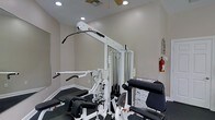 Located at 100 Carlton Park Dr in Flowood, Mississippi, this single-floor fitness facility offers a dedicated space for strength training and exercise. The room is designed with functionality in mind, featuring neutral-toned walls and a combination of recessed lighting and a ceiling fan that ensures the space remains well-lit and comfortable during workouts. The centerpiece of the facility is a Quantum multi-station cable and weight machine, a versatile piece of equipment that supports a wide range of strength training exercises including lat pulldowns, cable rows, and leg extensions. The machine is equipped with a weight stack system and multiple attachment points, making it suitable for users of varying fitness levels. A workout bench is also present, complementing the multi-station unit for added exercise variety. The flooring features a durable, textured surface appropriate for a fitness environment. Safety is accounted for with a fire extinguisher mounted on the wall. A reference chart is posted nearby, providing exercise guidance for users. The space is clean, organized, and purpose-built to support an effective fitness routine in a compact and practical setting.