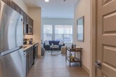 Located at 1850 Merchant Ln in Hampton, Virginia, this single-floor residence offers a thoughtfully designed layout with modern finishes and functional living spaces. The kitchen is a standout feature, outfitted with rich dark-toned cabinetry, granite countertops, a subway tile backsplash, and a full suite of stainless steel appliances including a refrigerator, dishwasher, electric range, and over-the-range microwave. Light wood-look flooring runs consistently throughout the unit, lending warmth and visual continuity to every room. The open living area is bright and welcoming, benefiting from large windows that bring in generous natural light and offer views of the surrounding community. The space accommodates both a seating and sleeping arrangement, making it highly versatile. A bathroom corridor connects to a dedicated laundry closet equipped with a stacked Whirlpool washer and dryer, providing the convenience of in-unit laundry. Additional storage is available via built-in shelving in the laundry area. The bathroom features granite-topped vanity cabinetry consistent with the kitchen's design language. Clean white walls, recessed lighting, and contemporary hardware complete the polished, move-in-ready aesthetic of this well-appointed Hampton residence.