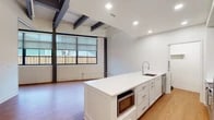 This single-floor space masterfully blends industrial character with contemporary design. The open living area showcases exposed steel beams and painted white brick walls, anchored by warm hardwood flooring that runs throughout. Large multi-pane windows line one wall, allowing abundant natural light to fill the space while offering views of the surrounding exterior.

The kitchen is a standout feature, presenting two distinct areas: an expansive white waterfall island with an integrated sink and built-in microwave, complemented by a secondary kitchen zone equipped with gray shaker-style cabinetry, subway tile backsplash, a stainless steel refrigerator, range with hood vent, dishwasher, and a stacked washer/dryer unit — a highly functional and well-appointed layout. A dedicated pantry or storage closet is accessible directly from the kitchen corridor.

The bathroom is finished to a refined standard, featuring full-height taupe tile surrounding the soaking tub and shower, a warm wood vanity with white quartz countertop, an under-mount sink, and a large backlit mirror. The tile flooring and accent wall tiling lend a cohesive, modern aesthetic throughout.

Overall, the space offers an inviting industrial-chic ambiance with thoughtful modern upgrades, making it both practical and visually striking.