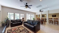 This single-floor space offers an open-concept layout that seamlessly connects the living and dining areas, creating a welcoming and functional environment. The living area is anchored by dark leather sofas arranged around a richly patterned area rug, providing a comfortable and stylish seating arrangement. Light-toned tile flooring extends throughout, lending a clean and cohesive aesthetic to the space.

The room is well-lit with recessed lighting complemented by two ceiling fans, ensuring both comfort and brightness across the expansive floor plan. Large windows and a glass-paneled door along the far wall invite generous amounts of natural light, enhancing the open and airy feel of the interior.

The dining area is positioned adjacent to the living space and features a solid wood dining table surrounded by matching chairs, ideal for shared meals or gatherings. A decorative niche with subtle accent lighting adds a touch of architectural character to the wall nearby.

Neutral-toned walls throughout the space provide a versatile backdrop that complements the warm wood furnishings and dark upholstery. The overall ambiance is casual yet polished, making this space well-suited for both everyday use and entertaining guests.