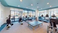 Located at 1588 Bloom St in Mount Pleasant, South Carolina, this modern fitness center is designed to support a comprehensive workout experience in a bright, welcoming environment. The single-floor facility is characterized by expansive floor-to-ceiling windows that line multiple walls, flooding the space with natural light and offering open views of the surrounding community.

The layout is thoughtfully organized into distinct workout zones. The cardio area is generously outfitted with a lineup of treadmills, elliptical machines, stationary bikes, and rowing equipment, all positioned to take advantage of the natural light and outdoor views. A wall-mounted television provides entertainment during workouts.

The strength training section features a variety of cable and resistance machines, along with a dedicated free weights area complete with a full dumbbell rack and adjustable weight bench. Full-length mirrors span an entire wall in this zone, enhancing the sense of space and allowing for proper form checks. Built-in open shelving runs below the mirrors, offering convenient storage.

The interior design reflects a coastal aesthetic, with a teal accent ceiling, white shiplap-style column details, and neutral-toned flooring throughout. Recessed lighting supplements the natural light, ensuring the space remains bright and energizing at any hour. A stretching table positioned centrally adds a functional recovery element to the facility.