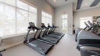 This single-floor fitness center is thoughtfully designed to accommodate a full range of workout needs across several distinct zones. The main strength training area features high ceilings accented by bold dark decorative beams with recessed lighting, creating a structured yet open atmosphere. Large mirrors line the walls, visually expanding the space while supporting proper form during exercise. The room is outfitted with a comprehensive selection of Cybex cable machines, functional training rigs with TRX attachments, adjustable benches, and a full dumbbell rack.

A dedicated cardio section houses a row of TRUE treadmills positioned along expansive windows that flood the area with natural light and offer views of lush tropical greenery. Additional cardio equipment includes a StairMaster stepmill and an Expresso stationary bike, situated in a bright, naturally lit corner. The flooring throughout is a durable, cushioned rubber-blend surface suited for high-impact activity.

A secondary open area provides ample space for stretching or functional movement, featuring a built-in storage nook with shelving and textured tile accent wall. Double doors with smart control panels offer convenient access points, completing this well-appointed facility.