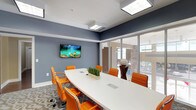 This contemporary conference room is thoughtfully designed to inspire productivity and creativity. At its center sits a large, sleek white oval conference table surrounded by vibrant orange ergonomic chairs, creating a bold and energizing color palette against cool blue-gray walls. The patterned carpet adds a layer of visual interest, grounding the space with a modern aesthetic.

A standout feature of the room is the full living green wall, densely covered in lush foliage and framing a natural light-filled window. This biophilic design element brings a refreshing, organic quality to the interior and serves as a striking focal point. Additional natural light filters in through large sliding glass partitions that open the space visually to an adjacent lounge or common area, enhancing the sense of openness.

Overhead, recessed skylight-style light fixtures provide bright, even illumination throughout the room. Wall-mounted flat-screen displays are positioned for clear visibility from all seating positions, making the space well-suited for presentations and collaborative meetings. Built-in power and data ports integrated directly into the conference table offer convenient connectivity.

Decorative touches, including colorful wall art and a small tabletop plant centerpiece, add warmth and personality to the space. The overall ambiance is professional yet inviting, balancing function with contemporary style.