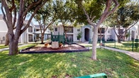 This outdoor playground reserve is a thoughtfully designed communal green space situated within a multi-building residential community. The centerpiece of the area is a green children's play structure set on a mulch-covered base, complete with slides, climbing elements, and activity panels, providing a safe and engaging environment for younger residents. Mature shade trees are distributed throughout the grounds, offering natural canopy coverage and a welcoming, park-like atmosphere on sunny days. Well-maintained grassy lawns extend across the space, bordered by clean concrete walkways that connect the various buildings and seating areas. Green metal benches are positioned along the paths, inviting residents to sit and enjoy the outdoors. The surrounding two-story residential buildings feature light blue and gray horizontal siding, arched entryways, black iron railings on upper-level balconies, and decorative landscaping including ornamental rocks and small shrubs. A green chain-link fence defines the boundary of the play area, adding a layer of safety. The overall ambiance is open, airy, and community-oriented, making this reserve a practical and pleasant outdoor amenity for residents of all ages.