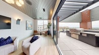 Located atop a building at Park Meadows Drive in Lone Tree, Colorado, this rooftop skydeck seamlessly blends indoor comfort with open-air living. The indoor lounge features light hardwood flooring, a distinctive slatted ceiling, and textured white walls that create a clean, contemporary aesthetic. Large floor-to-ceiling sliding glass doors connect the interior to the outdoor terrace, flooding the space with natural light and offering unobstructed views of the Lone Tree skyline and mountains beyond. The lounge is thoughtfully appointed with plush seating arrangements, wall-mounted flat-screen TVs, stylish wall sconces, and a long communal dining table beneath a modern chandelier — ideal for both casual gatherings and private events. The expansive outdoor terrace extends the entertaining space considerably, featuring a central fire pit lounge with generous seating, a bar-height dining area, and a built-in gas grill station. Warm timber accent panels and a partially covered pergola with string lights add ambiance to the outdoor area, while sleek glass-panel railings ensure uninterrupted panoramic views. Planter boxes with greenery and large-format tile flooring complete this elevated rooftop retreat.