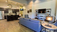 This well-appointed single-floor space offers an open-concept layout that seamlessly connects the living area and kitchen, creating a sense of spaciousness and flow throughout. The living area is anchored by a plush blue sofa and complemented by a sleek metal shelving unit, providing both comfort and functional display space. Soaring ceilings with white shiplap-style paneling draw the eye upward, amplifying the airy, open feel of the room. Rich hardwood floors run throughout, adding warmth and character to the space. A wall-mounted flat-screen television serves as a focal point in the living area, while an elegant chandelier with warm-toned lighting adds a touch of sophistication. The kitchen features dark cabinetry with a classic tile backsplash, built-in appliances, and a breakfast bar with seating, making it ideal for casual dining and entertaining. Accent walls in a soft yellow-green hue introduce a pop of color, contributing to the space's vibrant yet welcoming ambiance. Tasteful artwork and decorative accents are thoughtfully placed throughout, reflecting a curated, modern aesthetic that balances style with everyday livability.