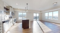 This single-floor unit presents a thoughtfully designed open-concept layout that seamlessly connects the kitchen and living areas. The kitchen is a standout feature, outfitted with gray shaker-style cabinetry, a white quartz countertop, a subway tile backsplash, stainless steel appliances including a range, microwave, and refrigerator, and a central island ideal for dining or food preparation. Pendant lighting above the island adds a modern touch, while light wood-tone flooring extends throughout the main living areas, enhancing the bright, airy atmosphere created by multiple large windows.

The living area is generously proportioned, bathed in natural light from expansive windows and a sliding glass door that opens to a private balcony overlooking the surrounding neighborhood. The bedroom is a comfortable, carpeted retreat featuring a large window with outdoor views and access to a spacious walk-in closet equipped with wire shelving systems. Two bathrooms are finished with light wood-tone flooring, white vanities with clean-lined cabinetry, and matte black hardware and towel bars, providing a cohesive modern aesthetic throughout. The overall ambiance is clean, contemporary, and light-filled.