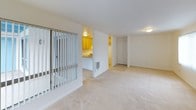 This single-floor residence is part of a well-kept community distinguished by its bold blue A-frame office building, mature palm trees, manicured lawns, and colorful agapanthus plantings. A winding concrete pathway leads through the landscaped grounds to a shared pool area enclosed by a white lattice fence, creating an inviting outdoor environment.

Inside, the unit features a practical, open layout with wall-to-wall carpeting throughout the living areas and bedrooms. The kitchen is equipped with warm honey-toned wood cabinetry, a double stainless steel sink set into a laminate countertop peninsula, and a freestanding electric range with overhead exhaust. The peninsula design opens the kitchen toward the living space, promoting a connected flow between cooking and living areas.

The living room is generously proportioned, featuring vertical blind-covered sliding glass doors that allow natural light to fill the room and provide views of the exterior. A closet is conveniently located off the main living area. The bedroom is spacious and bright, with a large window dressed in vertical blinds that filter natural light across the soft carpeted floor.

The bathroom features a tub and shower combination with full tile surround in a neutral cream palette, maintaining a clean and functional aesthetic throughout. Overall, the unit offers a comfortable, move-in ready living environment within an amenity-rich community setting.