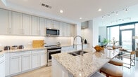 This thoughtfully designed single-floor residence offers a seamless blend of modern aesthetics and functional living. The open-concept kitchen serves as a centerpiece, showcasing crisp white shaker cabinetry extending to the ceiling, granite countertops with warm veining, a textured tile backsplash, and a full suite of stainless steel appliances including a French door refrigerator, electric range, and over-the-range microwave. A generous kitchen island with an undermount double sink and bar seating provides both workspace and casual dining options. Light wood-look flooring flows continuously throughout the space, enhancing the bright, airy atmosphere created by recessed lighting and a clean white palette. The living area offers an inviting layout with open sightlines that connect effortlessly to the kitchen. The bedroom is a standout, featuring floor-to-ceiling windows that frame sweeping panoramic city views and fill the room with abundant natural light, complemented by a ceiling fan. The primary bathroom is finished with granite countertops, white cabinetry, a framed vanity mirror, marble-look tile flooring, and a glass-enclosed shower with marble-style tile surround. A dedicated walk-in closet with built-in shelving is accessible directly from the bathroom, adding practical storage to this polished and cohesive residence.