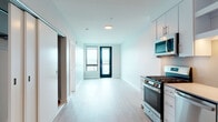 Located at 1211 W Columbia Way in Vancouver, Washington, this single-floor unit presents a clean, contemporary design throughout. The open-concept living and kitchen area is finished with light wood-tone flooring and bright white walls, creating a fresh, airy atmosphere that is further enhanced by natural light streaming through black-framed windows and a glass-panel exterior door leading to an outdoor space with expansive views of the surrounding landscape.

The kitchen is efficiently designed with white flat-front cabinetry, light-toned countertops, and a full suite of stainless steel appliances, including a gas range, over-the-range microwave, dishwasher, and refrigerator. Under-cabinet lighting adds a functional and polished touch to the workspace.

The bedroom is a well-proportioned, light-filled room with a large window framing outdoor views, complemented by the same wood-tone flooring found throughout the unit. A dedicated closet provides organized storage space.

The bathroom features full-height gray tile surrounding a soaking tub and shower combination, complemented by recessed lighting and wood-tone flooring that ties the space to the rest of the unit. A sliding door connects the bathroom and closet areas, contributing to the space's efficient and modern layout.