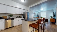 This single-floor residence offers a thoughtfully designed open-concept layout that seamlessly connects the kitchen, dining, and living areas. The kitchen serves as the centerpiece of the space, showcasing white shaker-style cabinetry extending to the ceiling, dark contrasting countertops, and a mosaic tile backsplash. A full suite of stainless steel appliances includes a French door refrigerator, range with oven, microwave, and dishwasher. A peninsula island with a dark countertop provides additional workspace and accommodates bar-height seating for casual dining.

Light hardwood-style flooring runs throughout the main living areas, contributing to a cohesive and airy feel. The living room benefits from natural light streaming through large windows with horizontal blinds, and features a teal accent wall that adds a pop of color to the otherwise neutral palette. A door from the living area provides additional access to the exterior.

The bedroom is generously sized with large windows that invite natural light, continuing the warm flooring and neutral tones. The bathroom features white cabinetry, a dark countertop vanity, a large mirror, and consistent flooring, maintaining the clean, modern aesthetic found throughout the space.