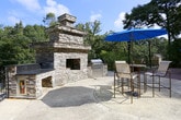 This expansive outdoor space offers a well-designed entertaining environment that seamlessly blends functional amenities with a natural, tree-lined setting. The main deck is constructed with warm-toned wood planking and is enclosed by black wrought iron railings, providing both safety and an unobstructed view of the surrounding greenery.

Multiple seating areas are distributed across the deck, including round dining tables paired with slatted metal chairs and large market umbrellas in bold blue, ideal for shaded outdoor meals. A dedicated lounging section features white chaise lounge chairs positioned to take full advantage of the open sky and lush canopy views.

Transitioning from the deck, a paved patio area serves as the focal point for outdoor gatherings, anchored by a striking stone fireplace and an adjacent built-in grill station. This area also accommodates additional dining with umbrella-shaded table seating, making it well-suited for both casual and social occasions.

The entire space is framed by mature deciduous trees that provide natural shade and a sense of privacy. The combination of the wood deck, stone hardscape, and verdant surroundings creates a cohesive and inviting outdoor retreat.