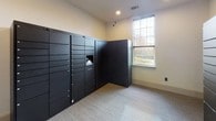 The package room is a single-level space designed to streamline parcel management through the use of Luxer One automated smart locker systems. Multiple freestanding locker units in a dark matte finish line the walls, arranged in an L-shaped configuration to maximize storage capacity. The lockers vary in compartment sizes, accommodating packages of different dimensions, from small envelopes to larger deliveries. A built-in digital interface on the primary Luxer One unit allows recipients to retrieve parcels securely using unique access codes. The room features neutral, warm-toned walls and low-pile patterned carpeting that contribute to a clean, functional aesthetic. Recessed ceiling lighting provides even illumination throughout the space. A window along one wall introduces natural light, adding to the room's brightness. Security cameras are positioned at upper wall corners, ensuring continuous monitoring of the area. The room connects directly to an adjacent corridor, allowing convenient access. Overall, the package room is thoughtfully designed to provide a secure, efficient, and well-organized environment for parcel handling.