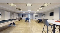 Located at 150 W Main St in Waukesha, Wisconsin, this generously sized single-floor amenity space is thoughtfully designed to accommodate both recreation and relaxation. The layout is divided into distinct functional zones that flow cohesively throughout the open floor plan.

The centerpiece of the space is a well-appointed game room featuring herringbone-patterned luxury vinyl plank flooring, a drop-panel ceiling with recessed lighting, and ample room for a pool table, foosball table, shuffleboard, ping-pong table, and arcade games. Soft blue-gray walls provide a clean, contemporary backdrop, while bold orange accent walls in the adjoining area add visual energy near the high-top seating and additional gaming zones.

A comfortable lounge area offers plush seating, patterned carpeting, and large windows that invite natural light and views of the surrounding grounds. An exterior door provides direct access to an outdoor courtyard. Adjacent to the lounge is a fully equipped kitchenette featuring stainless steel appliances, gray cabinetry, a marble-style backsplash, and wood-look flooring. A wall-mounted television and decorative artwork throughout contribute to the polished, inviting ambiance of this versatile community space.