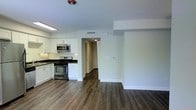 This single-floor space offers a clean, contemporary design with an open-concept layout that seamlessly connects the kitchen and living area. The kitchen is well-appointed with white shaker-style cabinetry, dark stone countertops, and a full suite of stainless steel appliances, including a gas range, over-the-range microwave, and additional cooking equipment. The crisp white cabinetry contrasts elegantly with the darker countertop surfaces, creating a polished and functional cooking space. Rich, dark hardwood-style flooring runs continuously throughout the unit, adding warmth and visual depth to the bright, neutral interior. The walls are finished in clean white tones, contributing to an open and airy ambiance. A hallway visible from the main living area leads to additional rooms, suggesting a practical and well-organized floor plan. Recessed lighting and overhead fixtures provide ample illumination, while HVAC vents integrated into the ceiling ensure year-round comfort. The overall design aesthetic is modern and move-in ready, combining durable materials with a neutral palette that offers a versatile backdrop for a variety of interior styles.