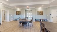 This 1230 square foot single-story community space offers a versatile and well-appointed environment for residents. The interior is characterized by light wood flooring throughout, creating a warm and inviting atmosphere. Recessed lighting and crown molding add a touch of sophistication to the space.

The layout includes a fitness center equipped with modern exercise machines, including treadmills, ellipticals, and strength training equipment. Large windows in this area allow for natural light and outdoor views.

A communal kitchen features dark wood cabinetry contrasting with light countertops and stainless steel appliances. Adjacent to the kitchen is a dining area with round wooden tables and chairs, perfect for gatherings or shared meals.

The space also includes office areas with desks, ergonomic chairs, and computer setups. A large decorative clock adorns one of the office walls, adding a stylish touch. Multiple seating areas throughout the space provide comfortable spots for relaxation or casual meetings.

French doors and large windows contribute to the bright, airy feel of the space, while neutral wall colors and tasteful artwork create a calming ambiance. The tray ceiling in the main area adds architectural interest and height to the room.

Overall, this community space combines functionality with aesthetics, offering residents a range of amenities in a contemporary and welcoming environment.