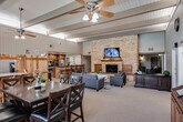 Located at 1540 W Ball Rd in Anaheim, California, this single-story community clubhouse offers a spacious, open-plan interior designed for gathering and relaxation. The layout flows seamlessly from a front reception area — framed by a distinctive wood-panel desk and roller shade windows that allow natural light — into a comfortable lounge zone featuring dark upholstered sofas, armchairs, and a plush area rug centered around a white coffee table. Exposed beam ceilings with ceiling fans lend architectural character throughout the space. A dramatic stacked-stone accent wall serves as the visual centerpiece of the main room, anchoring a wood-mantled fireplace topped by a wall-mounted flat-screen television. Adjacent to the fireplace, a formal dining area provides seating for a group around a dark wood table. The fully equipped kitchen is outfitted with rich wood cabinetry, granite countertops, stainless steel appliances including dual ranges and a refrigerator, and a stone-clad breakfast bar with bar stool seating — ideal for communal meals or events. A dedicated workstation area with computer terminals rounds out the functional offerings of the space. The warm, lodge-inspired design combines stone, wood, and neutral tones to create an inviting and well-appointed communal environment.
