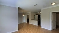 This single-floor space showcases an open-concept layout that seamlessly connects the living area to the kitchen, promoting a sense of spaciousness and ease of movement. The flooring throughout is hardwood-style planking in a warm natural tone, providing a cohesive and inviting foundation across the space.

The kitchen features a peninsula with a built-in sink, offering both additional counter space and a natural dividing point between the cooking and living areas. Dark cabinetry contrasts with the light countertops, while track lighting overhead ensures the kitchen remains well-illuminated. Open shelving is also visible, providing practical display or storage options.

Crown molding lines the ceilings, adding a refined architectural detail that elevates the overall finish of the space. The walls are painted in a soft, neutral gray-beige tone, contributing to a calm and versatile aesthetic that adapts well to a variety of furnishing styles.

A pendant light fixture is visible toward the rear of the space, hinting at a designated dining area. White interior doors and crisp baseboards complete the clean, polished look of this well-designed single-floor layout.