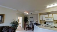 This single-story space showcases an open and connected layout that integrates the kitchen, dining, and living areas seamlessly. The dining area is anchored by a round table paired with ornate wooden chairs, positioned beneath a track lighting fixture that provides focused illumination. Crown molding lines the ceiling throughout, adding a refined architectural detail to the interior. A large decorative plant adds a natural, organic element to the space, complementing the warm, neutral color palette of the walls and carpet. Adjacent to the dining area, the kitchen features white upper cabinetry and a light-toned countertop that extends into a peninsula, creating a natural division between the kitchen and living spaces. A tray with candles and decorative accents sits atop the counter, contributing to the overall aesthetic. A framed botanical print on the wall adds a classic decorative touch. Through a doorway, an additional room is visible, suggesting a well-connected floor plan. The carpeted flooring and soft, warm lighting throughout give the space a comfortable and welcoming ambiance suited for everyday living.
