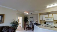 This single-story space showcases an open and connected layout that integrates the kitchen, dining, and living areas seamlessly. The dining area is anchored by a round table paired with ornate wooden chairs, positioned beneath a track lighting fixture that provides focused illumination. Crown molding lines the ceiling throughout, adding a refined architectural detail to the interior. A large decorative plant adds a natural, organic element to the space, complementing the warm, neutral color palette of the walls and carpet. Adjacent to the dining area, the kitchen features white upper cabinetry and a light-toned countertop that extends into a peninsula, creating a natural division between the kitchen and living spaces. A tray with candles and decorative accents sits atop the counter, contributing to the overall aesthetic. A framed botanical print on the wall adds a classic decorative touch. Through a doorway, an additional room is visible, suggesting a well-connected floor plan. The carpeted flooring and soft, warm lighting throughout give the space a comfortable and welcoming ambiance suited for everyday living.
