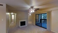 This single-floor space offers a well-appointed living area centered around a classic white-mantled fireplace, providing a focal point that adds warmth and character to the room. The ceiling features a textured finish and is equipped with a ceiling fan with integrated lighting, ensuring comfortable airflow and ambient illumination throughout the space. Soft, wall-to-wall carpeting extends across the living area, contributing to a cozy and comfortable atmosphere.

A sliding glass door on the far wall opens to an outdoor balcony, offering views of mature surrounding trees and creating a natural connection to the outdoors. The balcony, framed by a dark railing, allows natural light to filter into the interior during daylight hours.

A hallway extends from the living area, lined with built-in shelving and leading to additional rooms beyond. The walls are painted in a neutral tone, lending the space a clean and versatile aesthetic. Overall, the layout and features combine to create a functional and inviting environment suitable for comfortable everyday living.