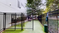 Located at 1305 Central Ave in Charlotte's Plaza Midwood neighborhood, this outdoor dog park offers a well-maintained, fully enclosed space designed for pet exercise and recreation. The park is surrounded by sturdy black metal fencing with multiple gated entry points, providing a secure environment for pets to roam freely. The ground is covered entirely in lush artificial turf, offering a clean and comfortable surface year-round regardless of weather conditions. Mature trees are positioned throughout the space, providing natural shade and a pleasant, park-like atmosphere. Wooden benches are placed along the perimeter, giving pet owners a comfortable spot to sit and supervise their animals. Waste management stations, including dispensers and receptacles, are conveniently located throughout the area to promote cleanliness and ease of use. Exterior wall-mounted lighting on the adjacent building ensures the space remains functional during early morning or evening hours. The surrounding landscaping, featuring neatly trimmed shrubs and mulched garden beds, adds to the overall aesthetic appeal of the area. This thoughtfully designed outdoor amenity combines functionality and comfort, making it a practical and enjoyable destination for pets and their owners alike.