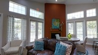Located at 100 Carlton Park Dr in Flowood, Mississippi, this single-floor space makes a striking first impression with its dramatic, soaring ceilings that lend an open and airy sense of grandeur. The main gathering area is anchored by a bold terracotta-toned accent wall that rises to the full height of the room, framing a classic wood-trimmed fireplace topped with curated décor, including a vibrant abstract canvas painting. Large, multi-pane windows line the walls on multiple sides, flooding the interior with generous natural light and offering pleasant views of the surrounding greenery outside. The high clerestory windows further enhance the sense of vertical space and brightness. The room is furnished with rich, dark leather seating accented by teal and coral throw pillows, creating a warm and inviting contrast against the neutral gray walls and light flooring. A dining area is visible nearby, complementing the lounge space and reinforcing the room's versatility for both relaxation and gathering. The overall ambiance is stylish yet comfortable, blending bold color choices with classic architectural details to create a space that feels both polished and welcoming.