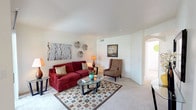 This single-floor space presents a comfortable and thoughtfully arranged living area that balances warmth with simplicity. The living room serves as the focal point, featuring light-colored carpeting that runs throughout, contributing to a clean and cohesive aesthetic. Smooth white walls and a flat ceiling create a bright, open atmosphere, complemented by a centrally positioned HVAC vent that ensures comfortable climate control year-round.

The room is well-proportioned, offering enough space to accommodate a full seating arrangement, including a rich red upholstered sofa adorned with textured accent pillows, a classic wingback armchair in warm taupe, and a glass-top coffee table resting atop a geometric patterned area rug. Decorative wall art and sculptural accents add personality to the space without overwhelming it. Ambient floor and table lamps provide warm supplemental lighting throughout the room.

A wide arched doorway leads to an adjacent area, suggesting a natural flow between living spaces. The overall ambiance is inviting and practical, with a neutral palette and warm accent tones creating a relaxed, livable environment suited for everyday comfort.