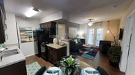 This single-floor residence offers a thoughtfully designed open-concept layout that seamlessly connects the kitchen, dining, and living areas into one cohesive and functional space. The kitchen is outfitted with dark-toned cabinetry, black appliances including a refrigerator and range, and a peninsula counter that serves as both a workspace and a casual divider between the kitchen and the rest of the living space. Adjacent to the kitchen, the dining area accommodates a full table set, decorated with tasteful centerpieces that add a welcoming touch. The living room is the centerpiece of the space, featuring warm hardwood-style flooring, a vibrant area rug with bold geometric patterns, and ample seating. A ceiling fan with a light fixture adds both practicality and style overhead. Natural light pours in through a large set of French doors fitted with blinds, offering an airy, open feel and a view of the outdoors. The accent wall in a warm gray tone adds depth and visual interest to the living area. The overall ambiance is modern yet comfortable, with a cohesive teal, gray, and neutral color palette that ties the entire space together elegantly.