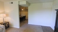 This single-story space showcases a thoughtfully arranged interior with a seamless flow between the living and dining areas. The living area features neutral-toned carpet flooring, while a transition to darker hardwood-style flooring is visible near the entryway, adding visual contrast to the space. Crisp white walls and white baseboards throughout create a clean, bright atmosphere.

A wide, open passageway with a structural overhead beam connects the living area to the formal dining room, providing a sense of defined yet open space. The dining room is carpeted and accommodates a dark wood dining table surrounded by matching dark upholstered chairs, making it well-suited for gatherings. A classic chandelier hangs above the dining table, providing warm, focused lighting. The dining room walls feature a subtle horizontal accent detail, adding a layer of architectural interest. The textured ceiling is consistent throughout both spaces. Overall, the interior presents a straightforward, functional layout with a neutral and welcoming ambiance.