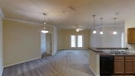 This single-story space showcases an open-concept layout that connects the kitchen and living areas into one cohesive environment. Crown molding runs along the upper perimeter of the rooms, adding a refined architectural detail throughout. The living area is finished with plush carpeting, while the transition zone near the kitchen features hard surface flooring, providing a practical and visually distinct separation between spaces. A ceiling fan is centrally positioned in the living area, promoting airflow and comfort. French doors at the far end of the living space introduce natural light and offer a view to the exterior. The kitchen area is partially visible and features dark countertops with pendant lighting hanging above, complementing an additional decorative pendant light near the entryway. Neutral-toned walls throughout contribute to a clean, understated aesthetic that allows the architectural details to stand out. A doorway leads to an adjacent room, suggesting additional private spaces beyond the main living area. Overall, the space presents a functional and well-appointed interior with classic design elements.