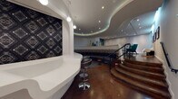 This sophisticated lounge space showcases a striking contemporary design defined by fluid, curved architectural forms throughout. The centerpiece is a dramatic sunken circular seating pit, lined with a continuous wraparound bench upholstered in deep navy, accented by a bold geometric patterned backrest in gold and blue tones. Small wooden stools and a patterned area rug complete the intimate gathering area. A sweeping staircase with dark wood treads and a sleek metal railing connects the sunken pit to the upper level, where teal upholstered lounge chairs and a white reception or service counter create a welcoming area. The counter is backed by a bold, dark diamond-patterned wall feature, illuminated by globe pendant lights. Layered, curved ceiling coffers with integrated LED accent lighting lend the space a dynamic, modern ambiance. Colorful glass vases serve as decorative accents along the upper ledge. A dedicated lounge nook on the upper level features teal armchairs arranged around a round white table, accompanied by framed black-and-white architectural photography and an artistic wall-mounted sculptural installation. Polished concrete flooring ties the areas together, reinforcing the space's modern, design-forward character.