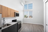 This contemporary single-floor studio showcases an open, airy layout defined by dramatically tall ceilings that amplify the sense of space throughout. The living area is bathed in natural light streaming through a stacked double-window configuration — a transom window above a standard casement — offering elevated views of the surrounding urban skyline and cityscape. Light-toned hardwood-style flooring runs continuously throughout, enhancing the bright, modern aesthetic.

The compact yet fully equipped kitchen lines one wall and features stainless steel appliances, including a range, built-in microwave, and refrigerator, complemented by warm wood-toned upper cabinetry, a dark tile backsplash, and dark countertops. A wall-mounted mini-split air conditioning unit provides efficient climate control.

A sliding barn-style door leads to the bathroom, which includes a vanity with an integrated sink, large mirror, and clean, neutral finishes. The unit also benefits from a private balcony accessible through floor-to-ceiling sliding glass doors, framed by decorative perforated metal railings and flanked by expansive windows that flood the space with light and provide open city views. The overall design is clean, minimalist, and well-suited to contemporary urban living.