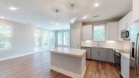This single-floor residence in Morrisville, NC offers a thoughtfully designed open-concept layout with contemporary finishes throughout. The kitchen is a standout feature, showcasing two-tone cabinetry with white upper cabinets and gray lower cabinets, a decorative arabesque tile backsplash, a center island with a bright white countertop, and a full suite of stainless steel appliances including a French door refrigerator, range, microwave, and dishwasher. Pendant lighting above the island adds a refined touch. The kitchen flows seamlessly into a generously sized living area bathed in natural light from multiple windows with views of surrounding greenery. Warm hardwood-style flooring extends throughout the main living areas, complementing the soft gray wall tones. The bedrooms feature plush carpeting and neutral walls, providing a calm retreat. The primary bathroom offers a white vanity with quartz countertop, hardwood-style flooring, and direct access to a walk-in closet equipped with multi-tier wire shelving. A second full bathroom includes a tiled tub-and-shower combination, a white vanity, and hardwood-style flooring. Recessed lighting throughout the space ensures consistent, well-distributed illumination in every room.
