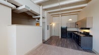 Located at 237 W Fifth Street in the heart of downtown Winston-Salem, North Carolina, this single-floor loft-style unit showcases an industrial-inspired design with striking architectural character. Soaring ceilings with exposed structural beams and visible ductwork define the open living and kitchen area, lending the space a distinctive urban aesthetic.

The kitchen is well-appointed with dark cabinetry, dark countertops, and a full suite of black appliances, including a refrigerator, electric range, and dishwasher. Ample upper and lower cabinet storage is complemented by a pendant light fixture that hangs from the impressive ceiling height above. Rich, dark hardwood-style flooring runs throughout the kitchen and main living area, creating a warm and cohesive look.

The carpeted bedroom area offers a quieter, softer retreat, with exposed piping along the upper walls that continue the loft-style industrial theme. The bathroom is clean and functional, featuring a walk-in shower stall with a curtain rod and a standard toilet, finished with hardwood-style flooring that ties back to the main living space.

A closet with double doors provides convenient storage near the entryway. Overall, this unit combines contemporary finishes with raw, industrial character, making it a compelling urban living option in Winston-Salem's vibrant downtown core.