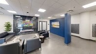 This contemporary lobby is a thoughtfully designed communal space that combines functionality with modern aesthetics. The open-concept layout is finished with large-format grey floor tiles and a suspended drop ceiling fitted with recessed panel lighting, ensuring the area is bright and evenly illuminated throughout.

Seating is arranged in multiple lounge groupings, featuring dark leather sofas and armchairs paired with round, low-profile coffee tables and a textured area rug — creating an inviting atmosphere for residents and guests. A striking bold blue accent wall serves as a visual centerpiece, adorned with a framed landscape artwork that adds a pop of color and character to the space.

A standout feature is a built-in corner aquarium filled with vibrant tropical fish, adding a lively and distinctive focal point near the entrance. The lobby also includes a prominently positioned mailroom bank with a large array of individual mailboxes, offering practical convenience for building occupants.

Architectural details include a prominent cylindrical structural column with a metallic finish, an electric fireplace set against a dramatic dark marble-effect wall panel, and glazed entry doors that invite natural light from the exterior. Potted plants and greenery are distributed throughout, adding warmth and a natural element to the clean, modern interior. The overall ambiance is polished, welcoming, and professionally finished.