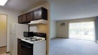 This single-floor space offers a straightforward, functional layout with an open connection between the kitchen and living area. The kitchen is equipped with a gas range, overhead range hood, and dark-stained upper cabinetry, providing ample storage. A countertop microwave adds convenience to the cooking space. The kitchen transitions seamlessly into the carpeted living area, which benefits from abundant natural light streaming through a large sliding glass door fitted with vertical blinds. The living room also features a wall-mounted air conditioning unit and a baseboard heating system, ensuring year-round comfort. The neutral wall tones and light-colored carpet throughout the living space contribute to a clean, understated ambiance. The sliding glass door offers views of exterior greenery, bringing a sense of openness to the interior. Overall, the space is designed with practicality in mind, making efficient use of its single-floor layout while maintaining a comfortable and livable environment.